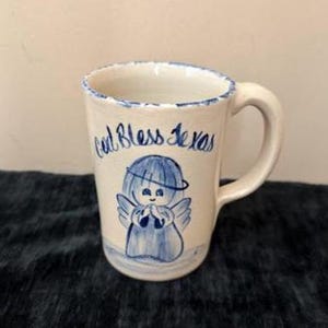 May include: A white ceramic mug with a blue rim and handle. The mug features a hand-painted angel design in blue, with the words "God Bless Texas" written in blue script. The mug is likely vintage or handmade.