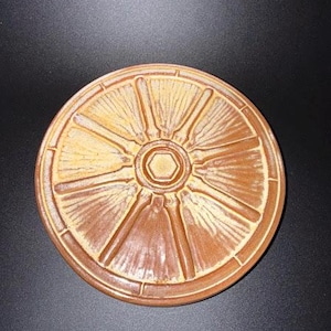 May include: A round, terracotta-colored ceramic coaster featuring a raised, embossed pattern resembling a citrus slice. The design includes a central hexagon surrounded by radiating spokes that divide the surface into segments, with a smooth, glazed finish.
