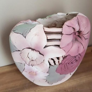 May include: A ceramic vase with a floral design. The vase is heart-shaped and features pink, white, and gray flowers. The interior of the vase is visible, with a series of horizontal ridges.
