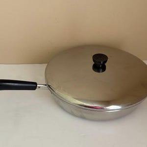 May include: A round stainless steel skillet with a polished finish, featuring a matching lid with a black knob handle. The pan includes a long, black heat-resistant handle extending from the side, which is equipped with a small metal hanging loop.