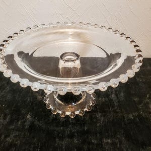 May include: A clear glass cake stand with a beaded edge and a central pedestal. The stand has a circular base and a raised, round platform. The glass is transparent, allowing light to pass through, and the beaded detailing adds a decorative touch.