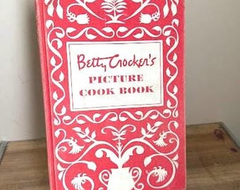 May include: A vintage cookbook titled "Betty Crocker's Picture Cook Book" with a red cover and white decorative scrollwork. The book's title is in a white rectangular box. The book is on a wooden surface.