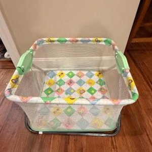 May include: A square portable playpen featuring mesh side walls and a padded floor mat with a colorful diamond pattern in shades of light blue, yellow, green, and pink. The frame is constructed with light green plastic corner connectors.