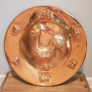 May include: A copper-colored decorative plate with a rooster in the center and bell designs around the rim. The plate has a shiny, reflective surface and is likely made of copper or a copper alloy. The rooster is in a standing pose.