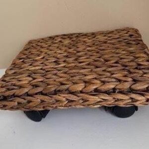 May include: A square plant caddy featuring a woven brown water hyacinth surface with a thick braided texture. The base is supported by four small black plastic swivel casters attached to the corners, allowing for mobility across flat surfaces.