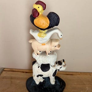 May include: A whimsical farm animal figurine featuring a rooster, goose, pig, sheep, and cow stacked on top of each other. The rooster has a red comb and yellow beak. The cow is white with black spots. The base is a dark green color.