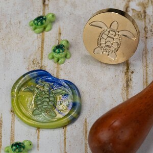 May include: A gold metal wax seal stamp with a turtle design. The stamp is on a white wooden surface with a blue and green wax seal and three small green turtle figurines.
