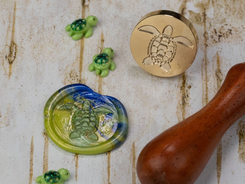 May include: A gold metal wax seal stamp with a turtle design. The stamp is on a white wooden surface with a blue and green wax seal and three small green turtle figurines.