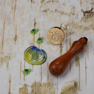 May include: A wooden handle wax seal stamp with a gold metal turtle design. The stamp is next to four small turtle figurines, one of which is a wax seal with a blue and yellow design.