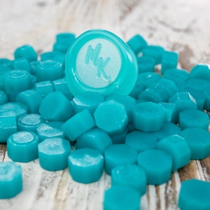 May include: A pile of teal blue wax seal stamps with one stamp already pressed with the initials "MK".