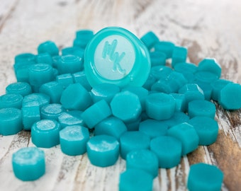 May include: A pile of teal blue wax seal stamps with one stamp already pressed with the initials "MK".