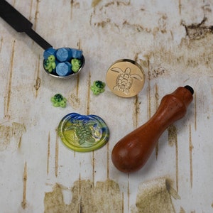 May include: A set of wax seal supplies featuring a wooden handle, a gold metal seal with a turtle design, and a small spoon filled with blue and green wax beads. There are also several small turtle charms.