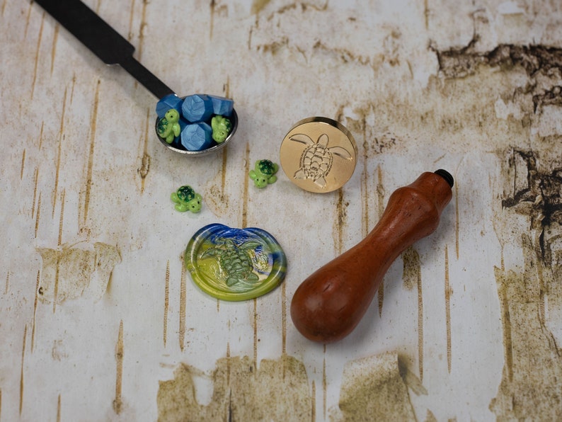 May include: A set of wax seal supplies featuring a wooden handle, a gold metal seal with a turtle design, and a small spoon filled with blue and green wax beads. There are also several small turtle charms.
