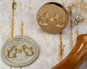 May include: A gold-toned metal stamp with two cats facing each other and a heart between them. The stamp is used to create wax seals. A wax seal with the same design is shown next to the stamp.