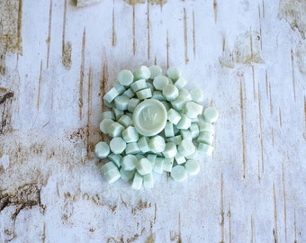 May include: A collection of pale green wax seals, some scattered and some in a pile, with one seal bearing the initials "ML". The seals are on a textured, light-colored wooden surface.