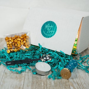 Può includere: Una scatola bianca con un logo turchese che dice "Waxing Poetic" e un sigillo turchese con il testo "sigilli di cera personalizzati" è aperta e riempita di carta crespa turchese. La scatola contiene un sacchetto di perline di cera dorate, un timbro per sigilli di cera turchese, un cucchiaio di metallo turchese e una candela da tè bianca.