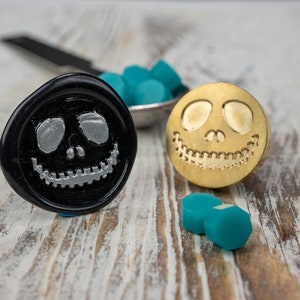 May include: Two wax seal stamps with a smiling skull design. One stamp is gold and the other is black with a silver skull. There are several teal wax seals scattered around the stamps.