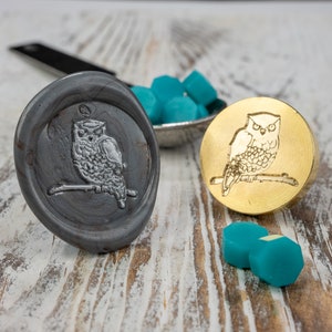 May include: Two wax seals with an owl design. One seal is gold and the other is gray. Both seals are on a white wooden surface. There are teal wax beads in the background.