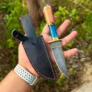 May include: A fixed-blade knife featuring a Damascus steel blade with a wavy pattern, a brass bolster, and a handle crafted from a combination of light brown wood and translucent blue resin, accompanied by a black leather sheath with yellow stitching.