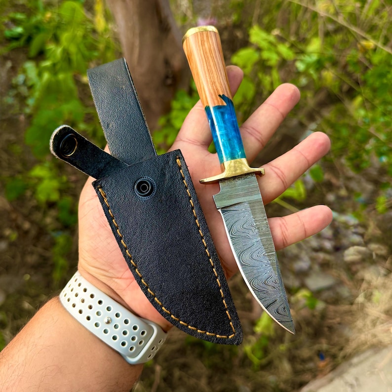 May include: A fixed-blade knife featuring a Damascus steel blade with a wavy pattern, a brass bolster, and a handle crafted from a combination of light brown wood and translucent blue resin, accompanied by a black leather sheath with yellow stitching.