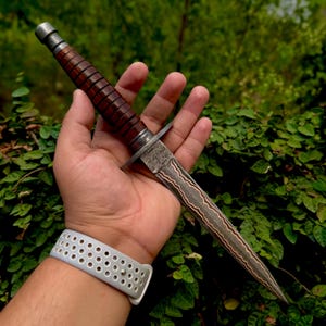 May include: A double-edged dagger featuring a dark brown, ribbed cylindrical handle with a metallic pommel and a crossguard. The blade displays a distinct wavy, layered pattern along its length, transitioning from a textured bolster to a sharp point.