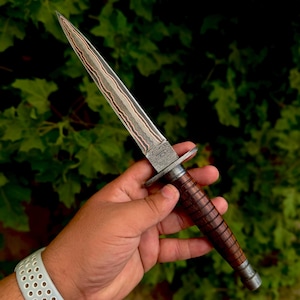 May include: A double-edged dagger featuring a pointed blade with a visible layered Damascus steel pattern. The handle is constructed from dark brown wood with a series of horizontal grooves and a metal crossguard separating the blade and grip.