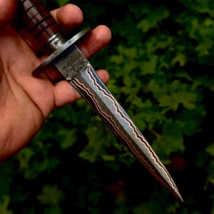May include: A double-edged dagger featuring a blade with a distinct wavy pattern of layered steel and copper. The handle is constructed from dark brown, grooved wood, separated from the blade by a polished metal guard with a textured finish.
