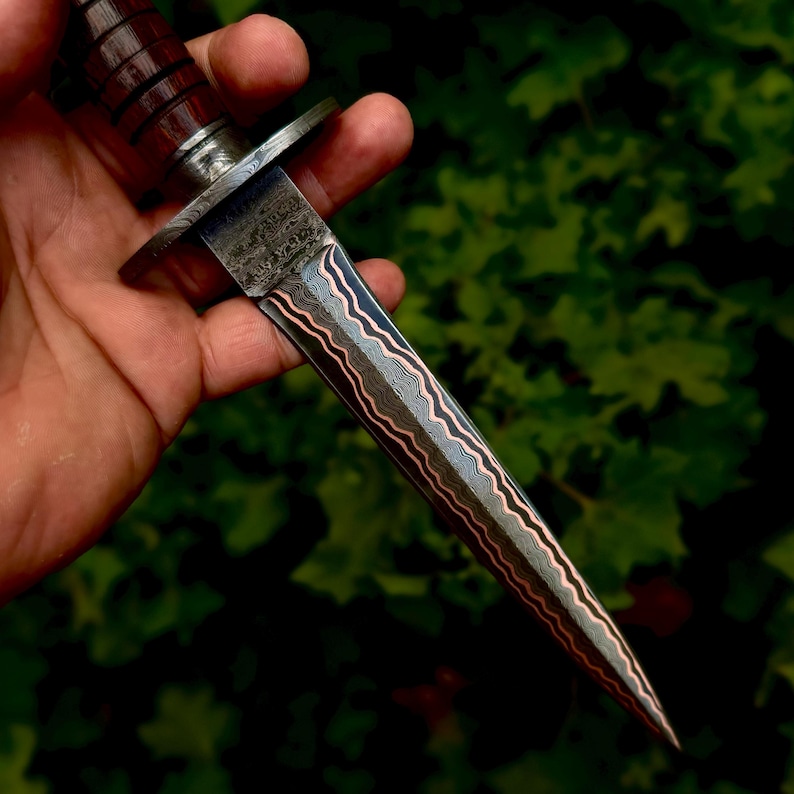 May include: A double-edged dagger featuring a blade with a distinct wavy pattern of layered steel and copper. The handle is constructed from dark brown, grooved wood, separated from the blade by a polished metal guard with a textured finish.