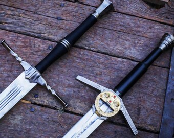 May include: Two swords with black handles and silver blades. One sword has a decorative gold pommel with red accents. The swords are lying on a wooden surface.
