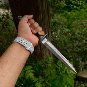 May include: A double-edged dagger with a patterned Damascus steel blade featuring a copper-colored center line. The handle is dark wood with a metal guard and pommel. A light gray silicone watch band with a perforated design is visible on the wrist.