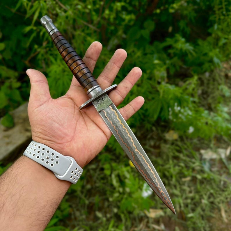 May include: A double-edged dagger featuring a dark brown, ribbed wooden handle and a metal crossguard. The blade displays a distinct wavy, layered pattern with a copper-colored center line running down the length of the sharp, pointed steel edge.