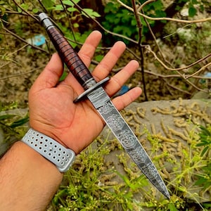 May include: A fixed-blade knife featuring a dark brown, ribbed wooden handle and a silver-toned metal crossguard. The blade displays a distinct, swirling Damascus steel pattern that extends from the bolster to the sharp, pointed tip.