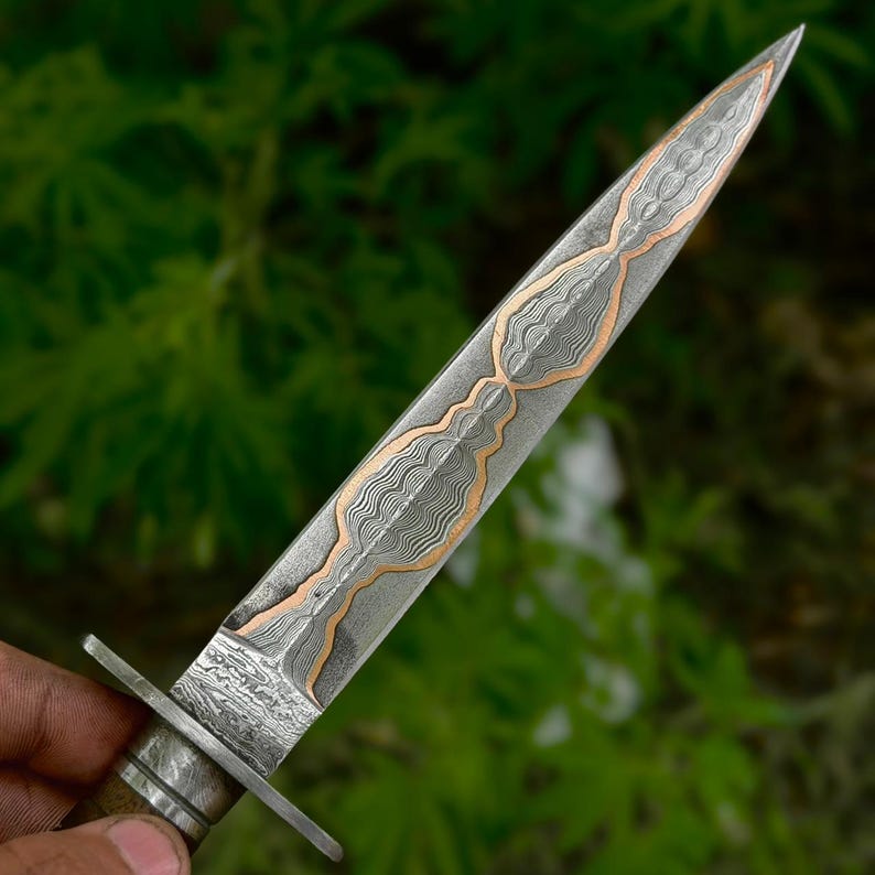 May include: A double-edged dagger featuring a pointed blade with a complex, wavy Damascus steel pattern integrated with a central copper inlay. The blade has a distinct metal guard separating it from the handle, showcasing intricate metalwork.