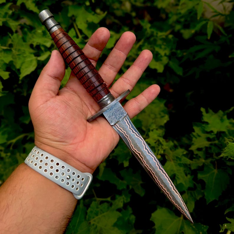 May include: A double-edged dagger featuring a dark brown, ribbed wooden handle with a metallic pommel and a crossguard. The blade displays a distinct wavy, layered pattern along its length, tapering to a sharp point from the central spine.