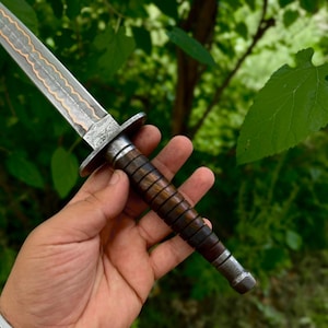May include: A dagger featuring a patterned Damascus steel blade with a central copper inlay, a metal crossguard, and a dark brown wooden handle with horizontal grooves, finished with a metal pommel at the base of the grip.