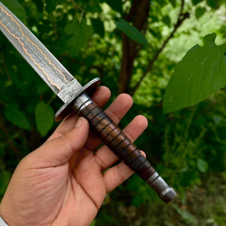 May include: A dagger featuring a patterned Damascus steel blade with a central copper inlay, a metal crossguard, and a dark brown wooden handle with horizontal grooves, finished with a metal pommel at the base of the grip.