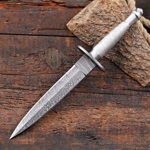 May include: A double-edged dagger featuring a pointed blade with a distinct wavy Damascus steel pattern. The handle is made of silver-toned metal with a textured, cross-hatched grip and a rounded pommel, separated from the blade by a flat guard.