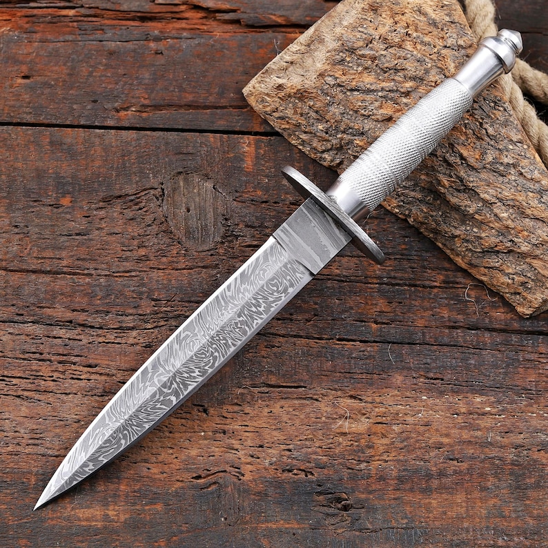 May include: A double-edged dagger featuring a pointed blade with a distinct wavy Damascus steel pattern. The handle is made of silver-toned metal with a textured, cross-hatched grip and a rounded pommel, separated from the blade by a flat guard.