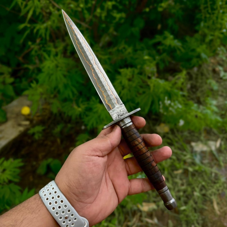 May include: A double-edged dagger featuring a pointed steel blade with a central copper-colored wavy inlay. The handle is constructed from dark brown wood with horizontal grooves and a silver-toned metal crossguard and pommel cap at the base.