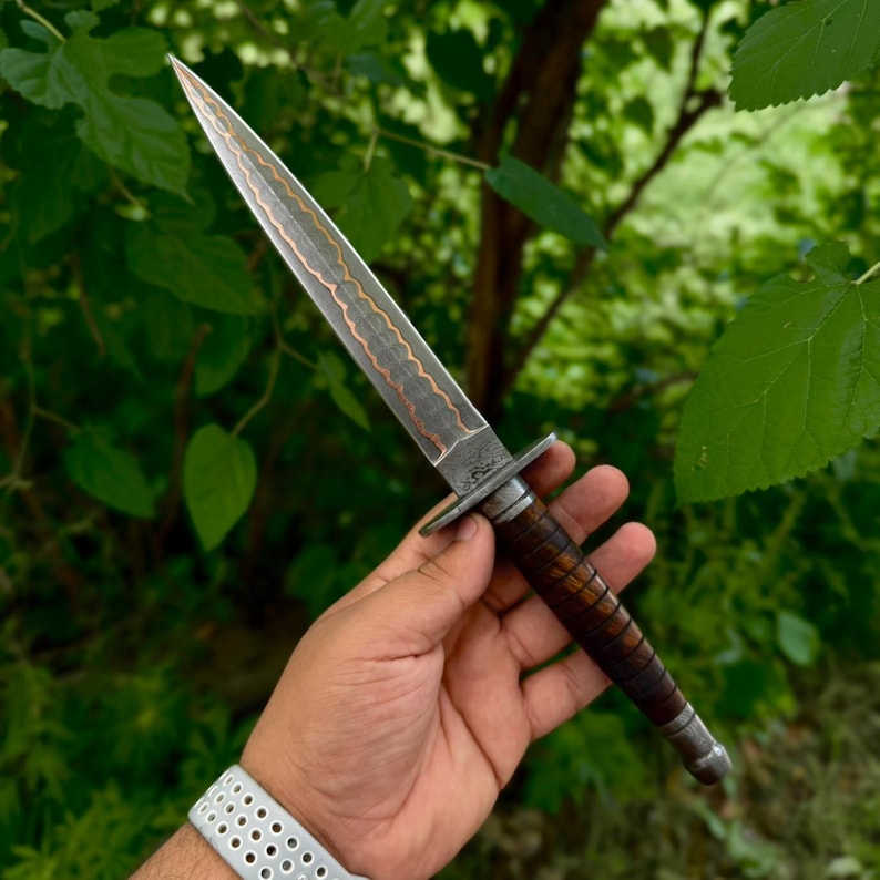 May include: A double-edged dagger featuring a pointed steel blade with a central wavy copper inlay. The handle is constructed from dark brown stacked leather rings, finished with a steel crossguard and a rounded pommel at the base of the grip.