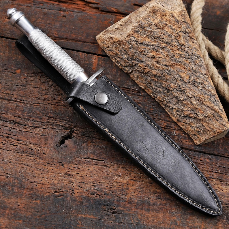 May include: A silver-colored metal knife with a textured, knurled handle and a pointed tip, partially inserted into a black leather sheath. The sheath features visible light-colored stitching along the edges and a single metal snap button closure.
