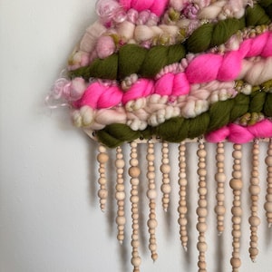 May include: A handmade wall hanging featuring a woven design with bright pink, olive green, and cream-colored yarn. Wooden beads hang from the bottom. The textile art piece is mounted on a wooden base and is displayed against a white wall.