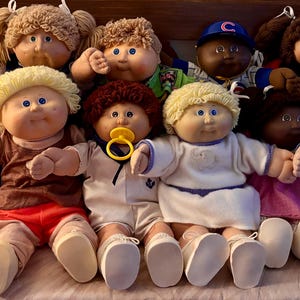 May include: A collection of Cabbage Patch Kids dolls, featuring various skin tones, hair colors, and outfits. Some dolls wear baseball caps, while others have pacifiers. The dolls are arranged in a group, showcasing their diverse appearances.