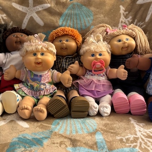 May include: A collection of seven Cabbage Patch Kids dolls with diverse hair colors, skin tones, and outfits. The dolls feature signature round faces, yarn hair, and fabric bodies, with one doll wearing a pink pacifier and another wearing an ABC print shirt.