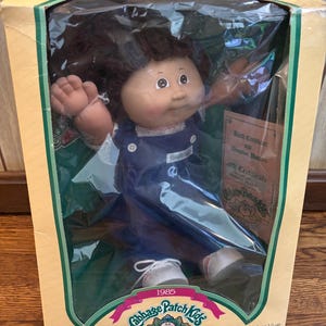 May include: A Cabbage Patch Kids doll in its original packaging. The doll has brown hair, a fair complexion, and is wearing a blue dress and white shoes. The box features the Cabbage Patch Kids logo and the text "1985".