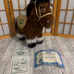 May include: A brown plush horse toy with a black mane and tail, wearing a tan saddle. The toy has white fur on its hooves and a gold-colored bridle. Accompanying the toy are two printed documents.