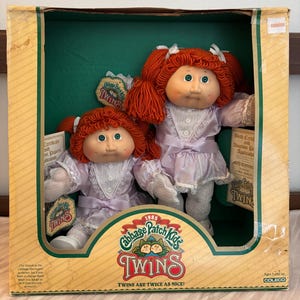 May include: A vintage Cabbage Patch Kids Twins doll set in its original box. The dolls have red yarn hair, green eyes, and wear lavender dresses. The box features the Cabbage Patch Kids logo and the text "Twins are twice as nice!"