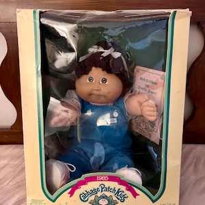 May include: A 1985 Cabbage Patch Kids doll with dark brown yarn hair and a white ribbon, wearing blue overalls over a white shirt. The doll is inside its original packaging, which includes a Birth Certificate and Adoption Papers document.