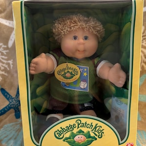 May include: A Cabbage Patch Kids doll with light brown curly hair and blue eyes, wearing a green and brown sports jersey. The doll is packaged in a yellow and green cardboard box with the text Cabbage Patch Kids and One of a Kind. And Just Mine!
