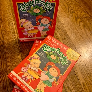 May include: A red box of 32 Cabbage Patch Kids Valentines, with two additional boxes visible. The boxes feature illustrations of Cabbage Patch Kids and text that reads "Includes 2 Cards on Back of Box" and "Free Coupon Inside".